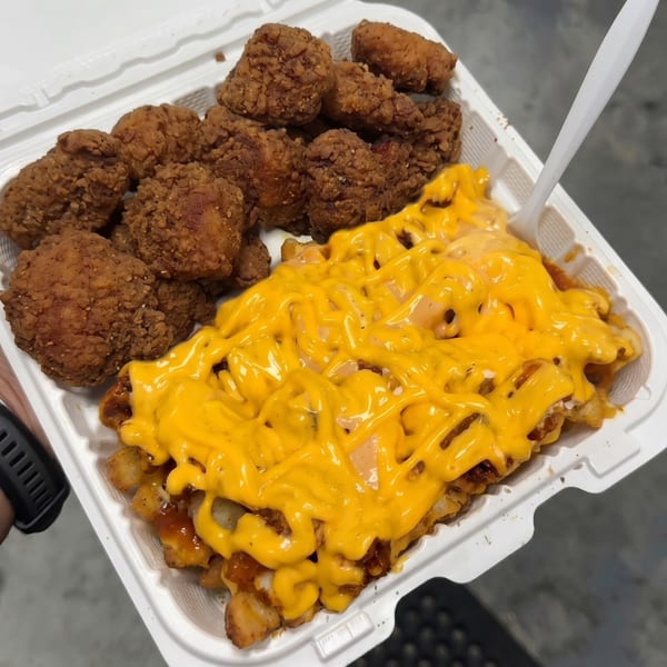 A white takeout container holds crispy fried chicken nuggets and waffle fries smothered in cheese sauce.